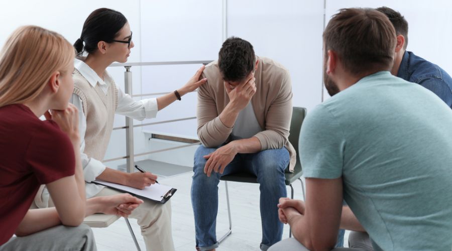 A group therapy session at a treatment facility shows five people seated in a circle. One man appears distressed, while a woman beside him offers comfort with a hand on his shoulder, reflecting the supportive environment of IOP programs.
